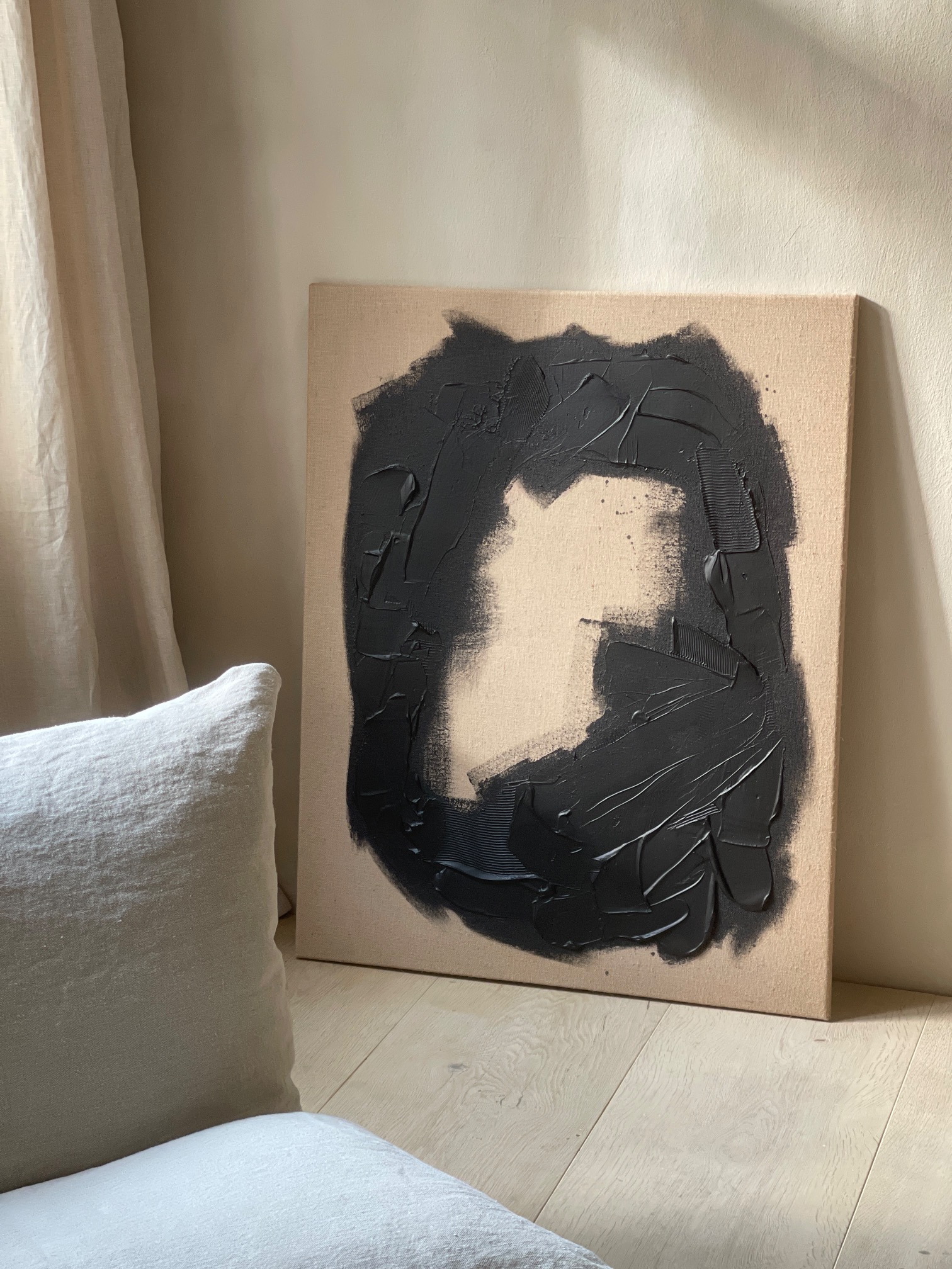 Acrylic painting with black abstract motif on jute canvas stands on a light-colored floorboard, with a lounge chair in front of it in the section